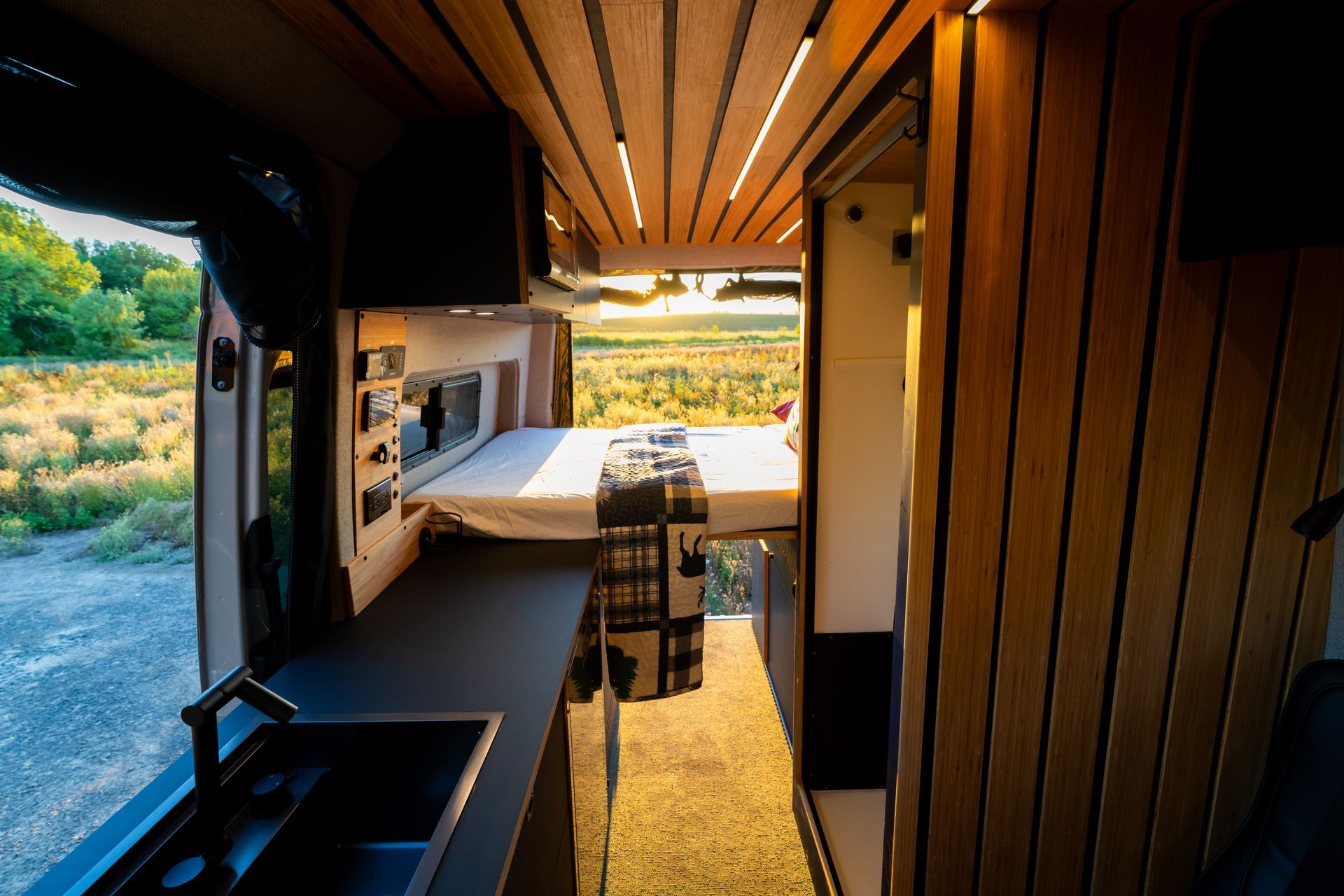 Modern camper van interior with bed, kitchenette, and open view to a sunny outdoor landscape