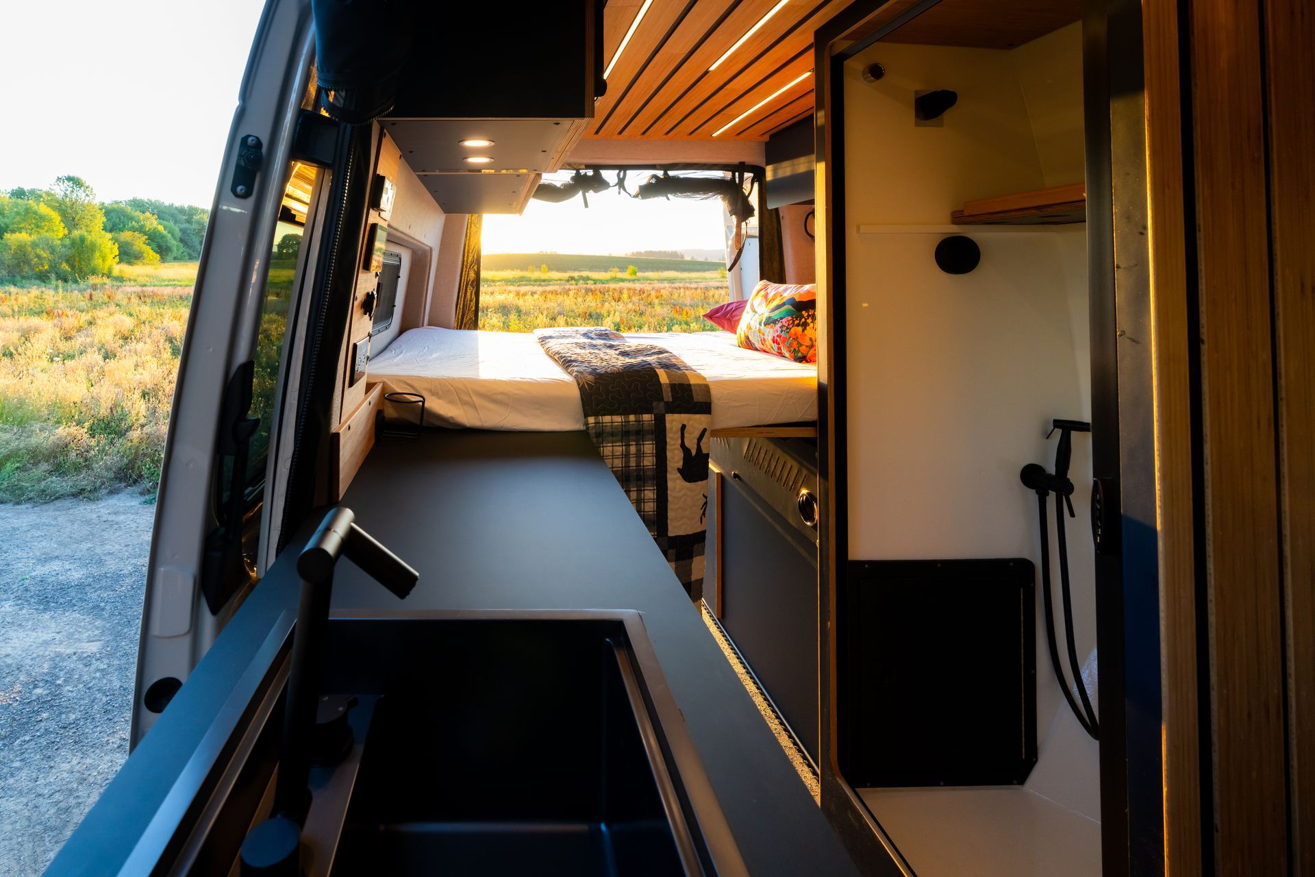 Compact camper van interior with a kitchenette, bench seating, and open door overlooking a grassy field