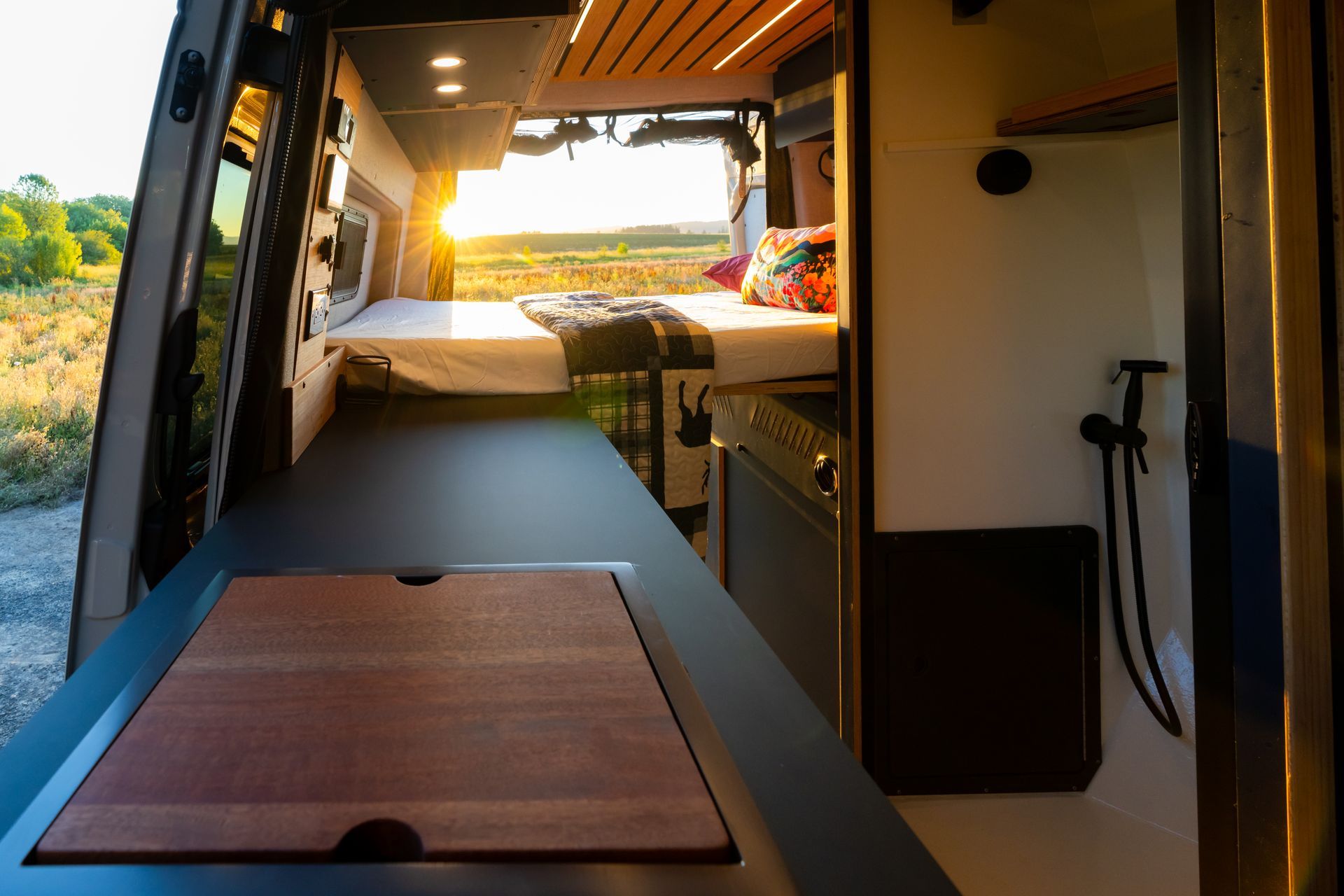 Modern camper van interior with a bed, wooden table, and sunset view through the open rear door