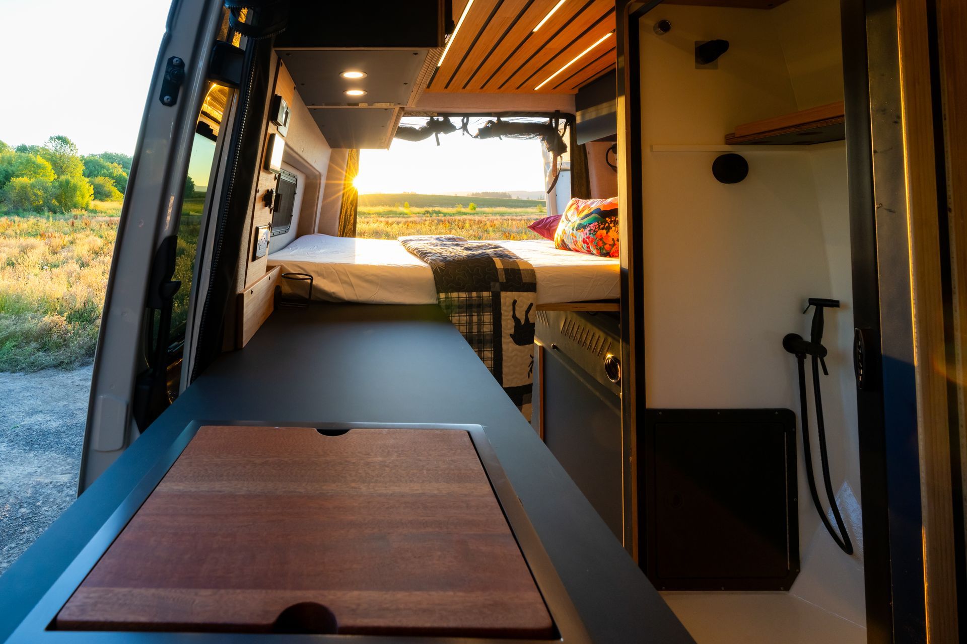 Inside a camper van: bed, wooden countertop, sink, and open sliding door with a view outside.