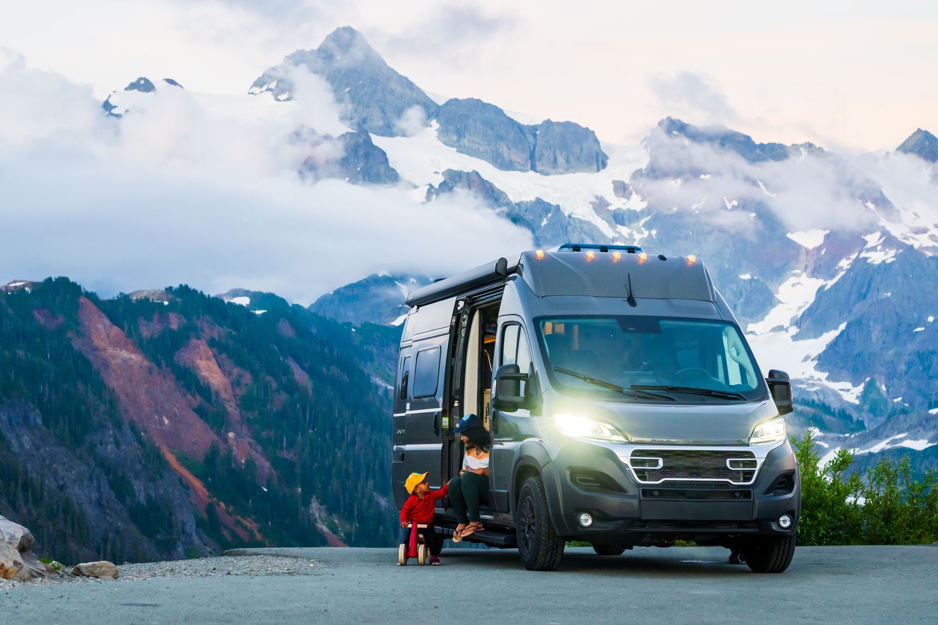 Gray RV on a mountain road with headlights on, next to a person in red jacket, snowy peaks behind