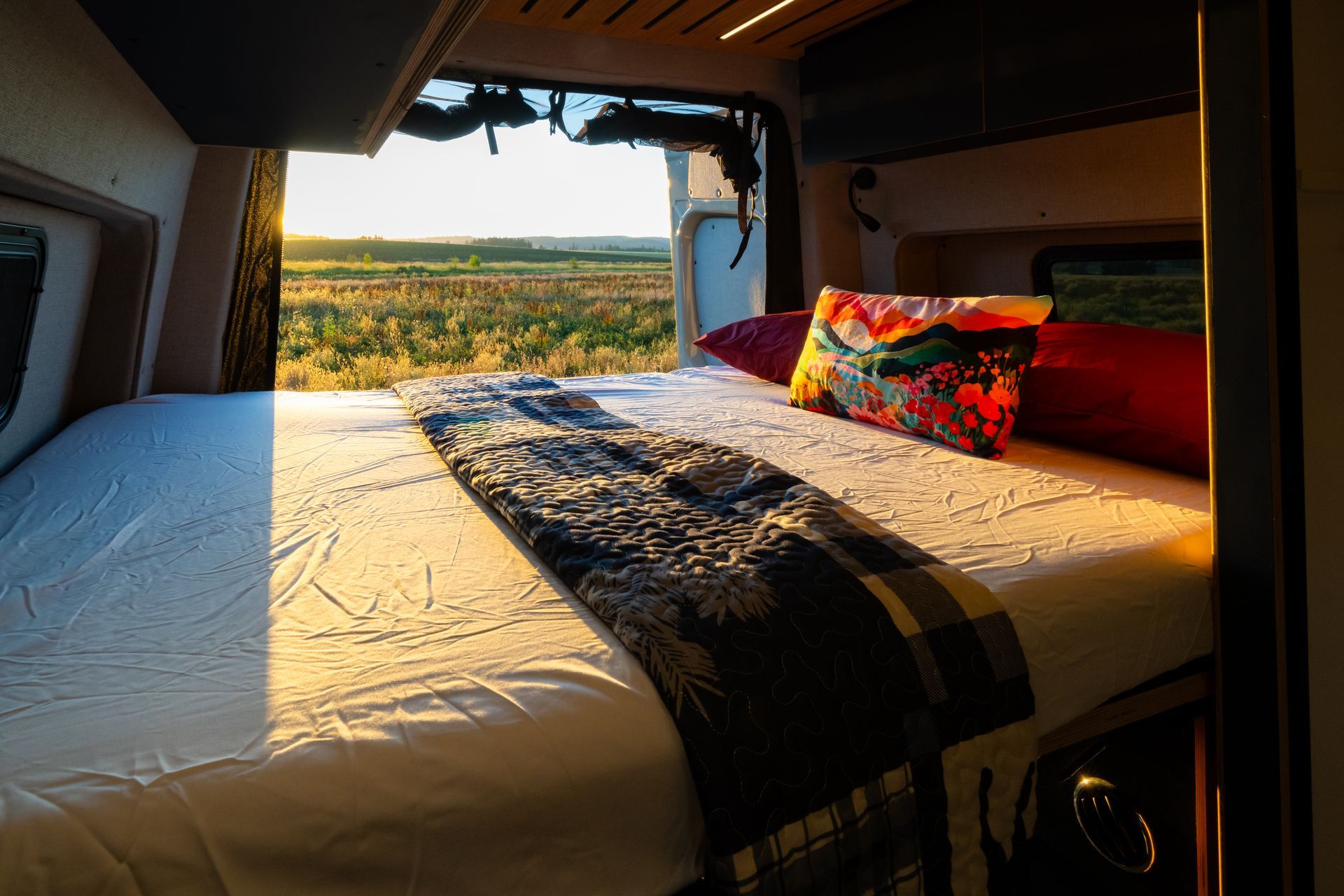 Bed in a camper van by an open door, with sunset view over fields and colorful pillows