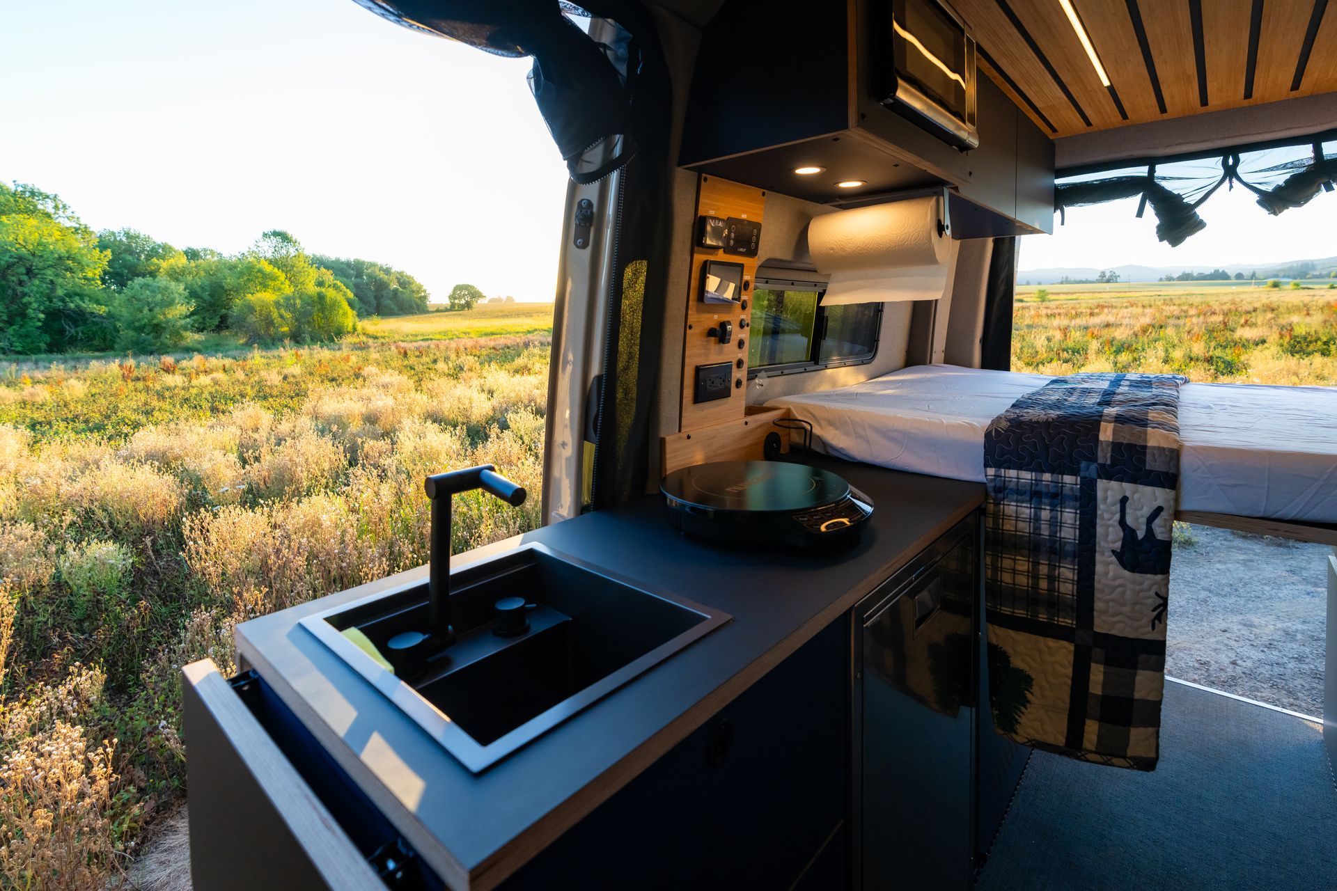 Cozy camper interior with bed and kitchenette overlooking a sunny grassy field