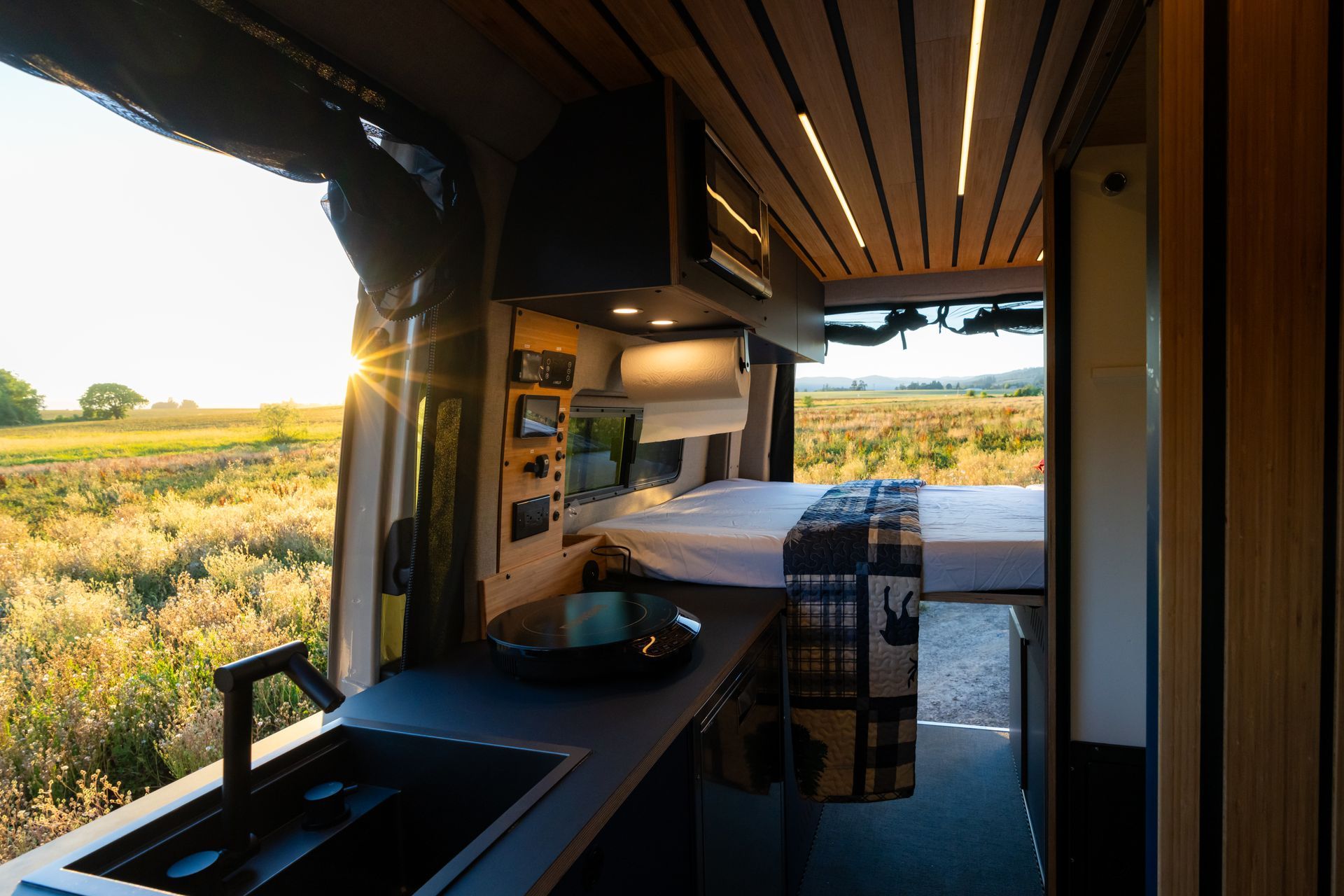Modern van interior with a bed and desk, looking out over a sunlit grassy landscape.