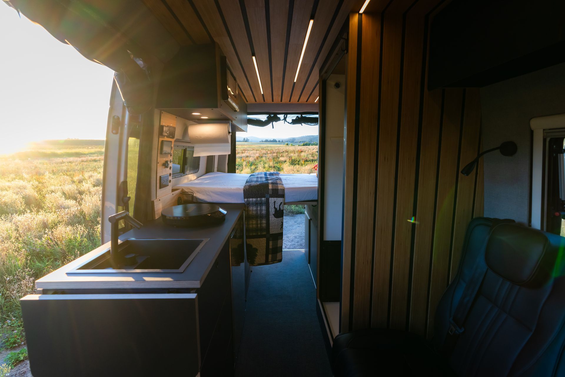 Modern RV interior with kitchen, wood-paneled walls, and open door overlooking a sunny landscape
