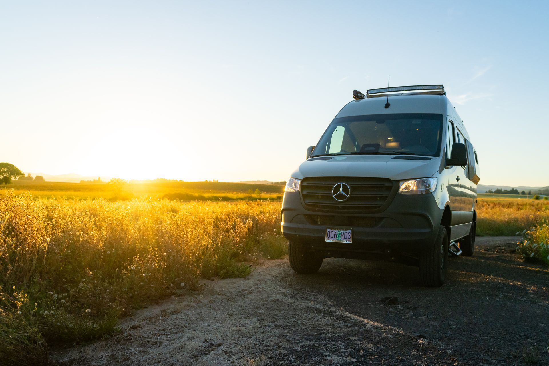 Black camper van on a gravel road beside a golden field at sunset.