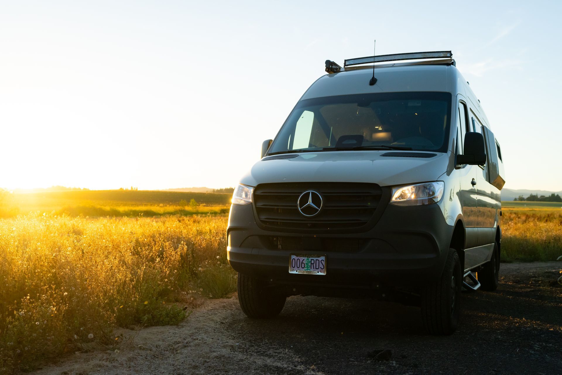 Black van parked on a dirt road beside a sunlit field at sunset