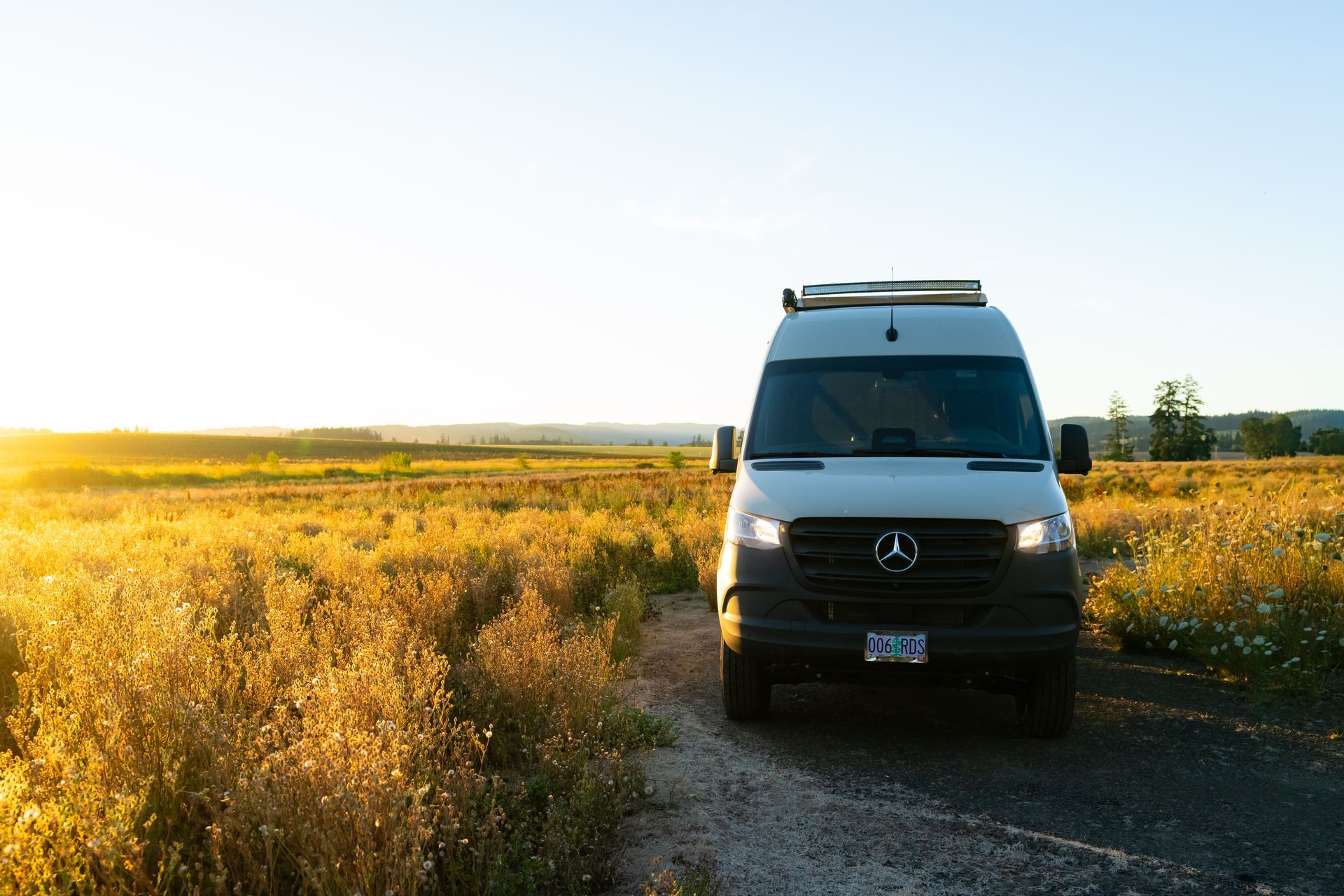 White van driving on a dirt road beside a golden field at sunset