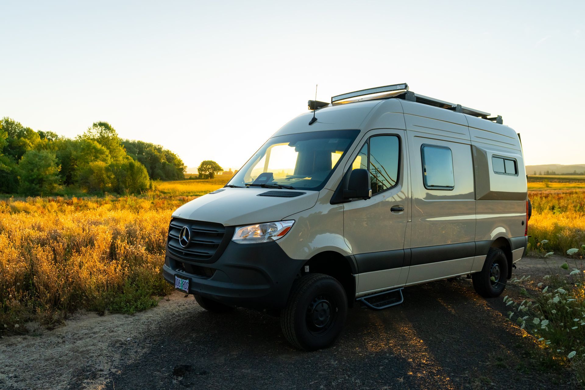 Silver camper van parked on a dirt path beside a golden field at sunset