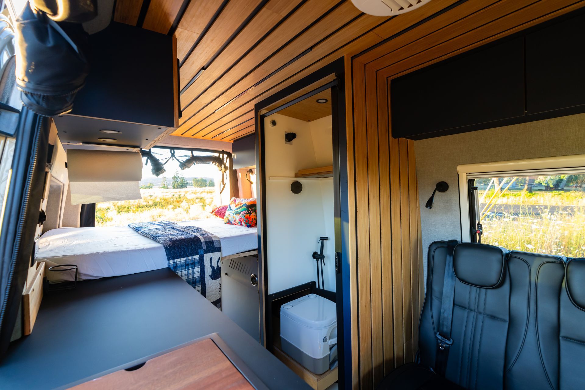 Interior of a camper van with wood ceiling, bed, and black seat by the open side door.