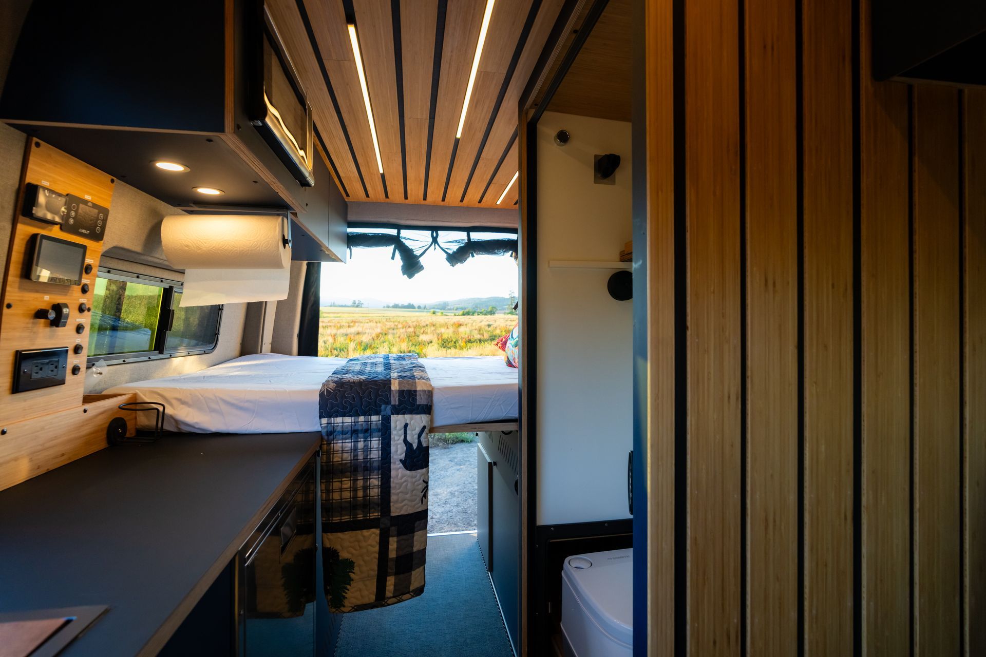 Compact camper van interior with bed, wooden slats, and open rear doors overlooking a scenic landscape