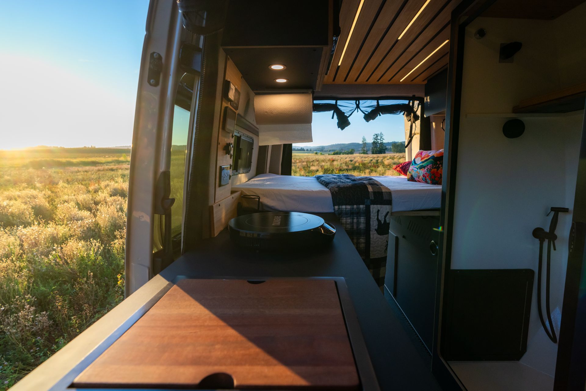 Open camper van interior with table, bed, and sunset view over a grassy landscape