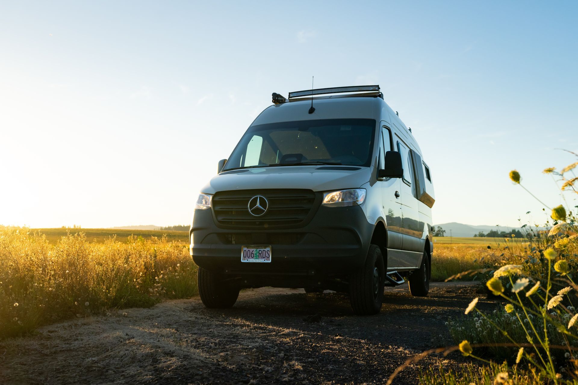 White camper van parked on a dirt road beside a sunlit field at sunset