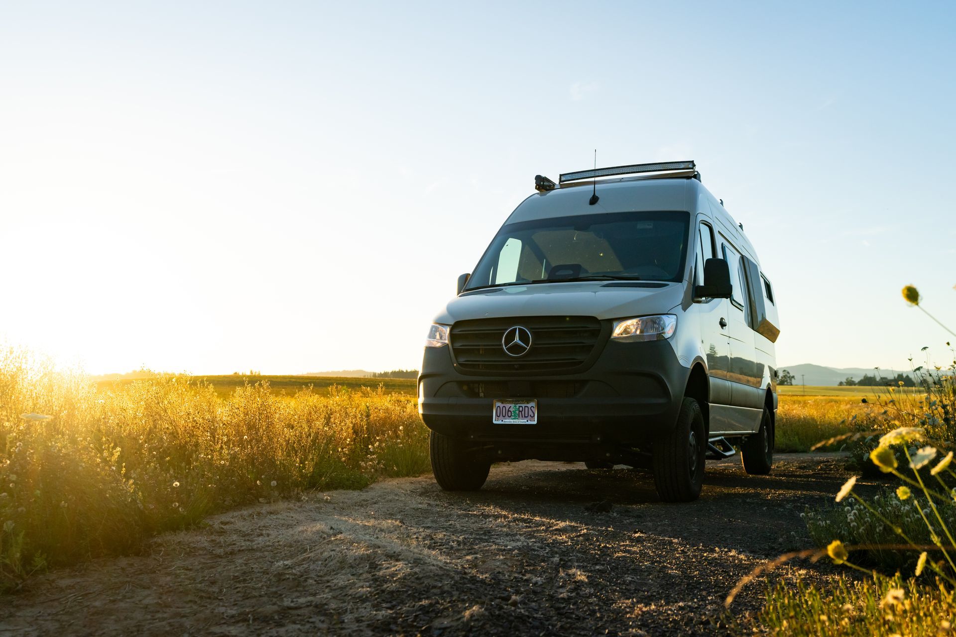 Silver camper van on a dirt road beside a sunlit field at sunset