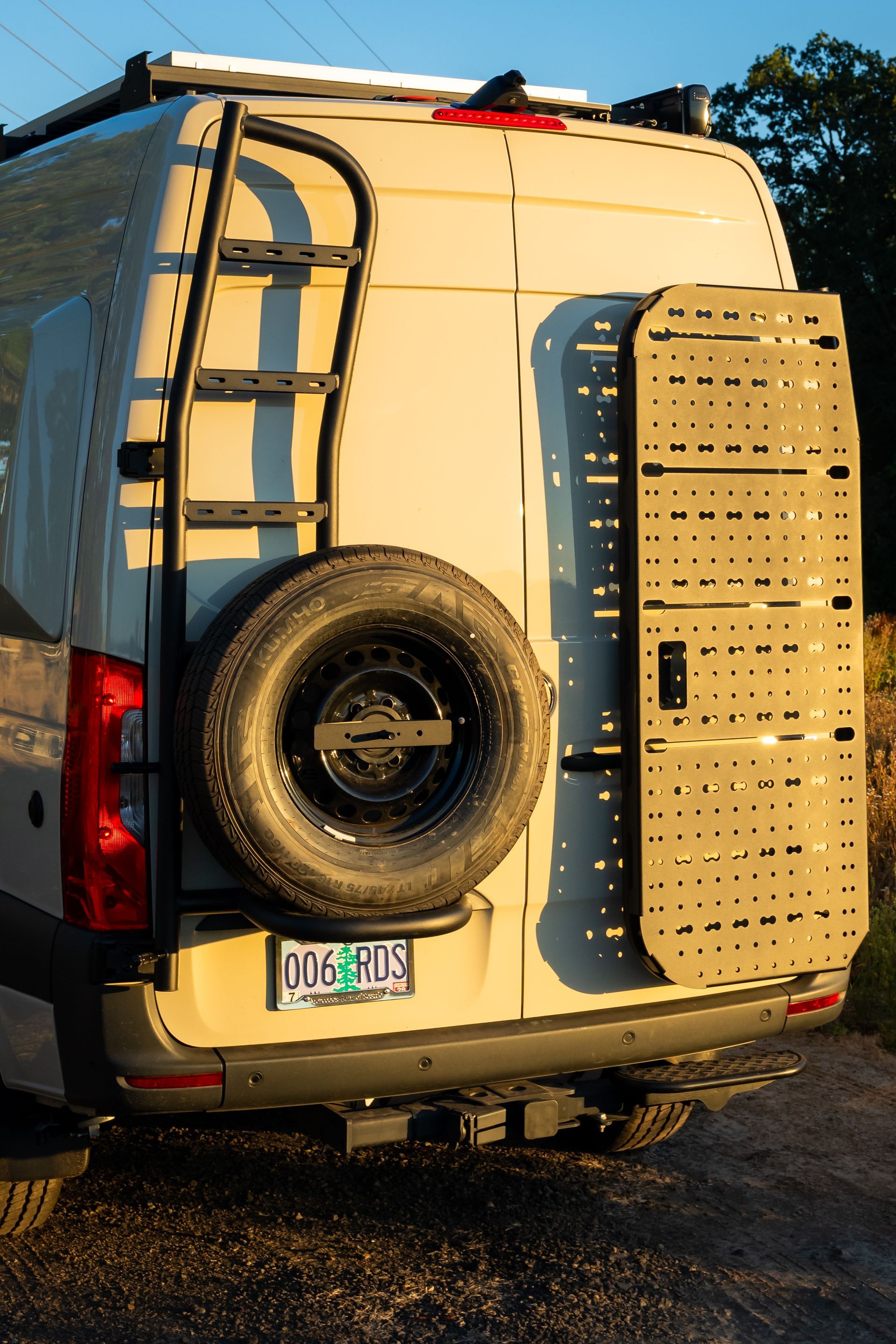 White van rear with spare tire and ladder on back, parked on a gravel road at sunset.