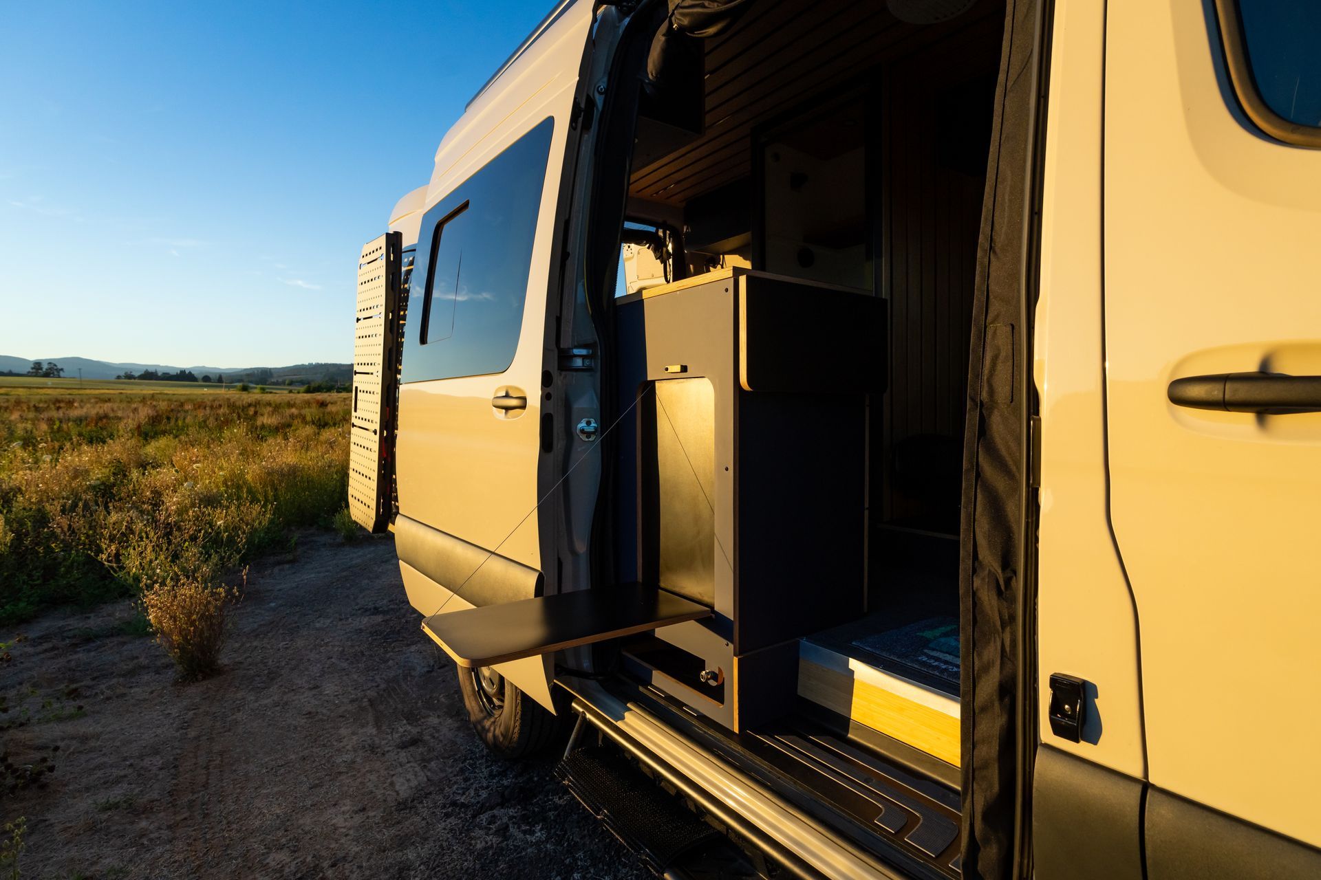 Open van parked on a rural road with its side door open beside a grassy field at sunset