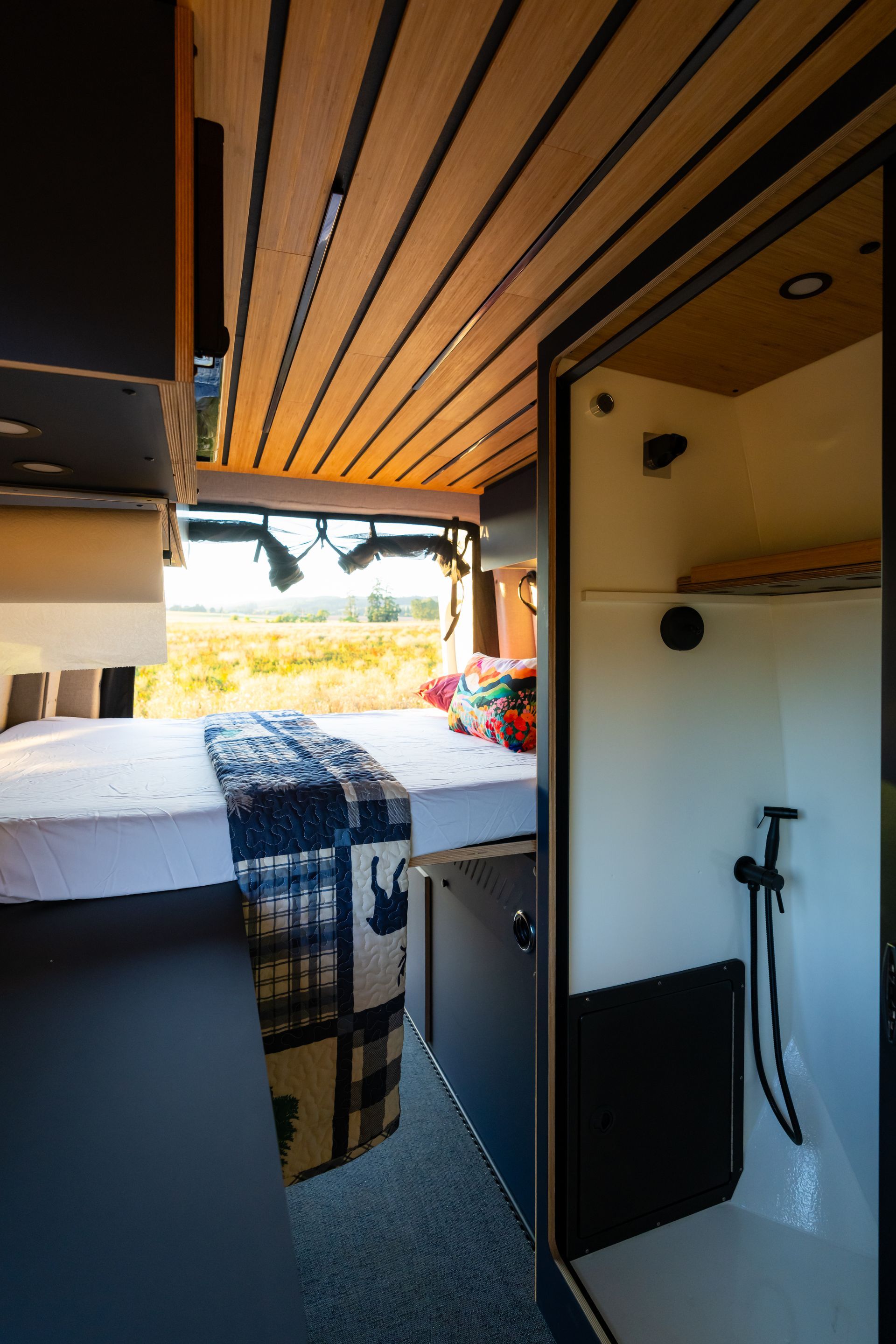 Compact camper interior with bed, open side door, white shower stall, and slatted wooden ceiling