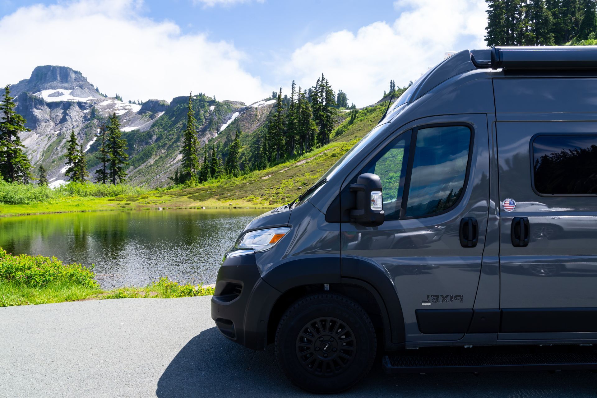 Dark camper van parked by a mountain lake with snowy peaks and pine trees in the background