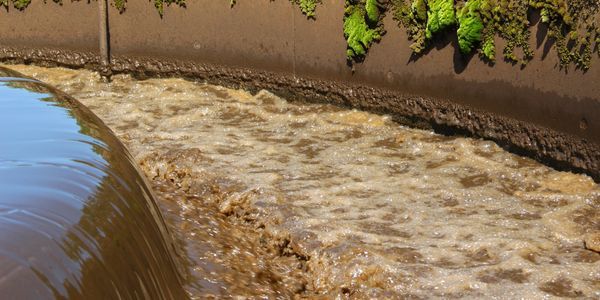 Murky water rushing through a concrete canal beside a brown embankment with green vegetation.