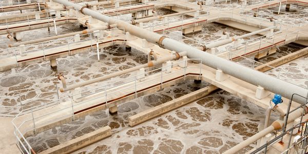 Aerial view of a wastewater treatment plant with beige tanks, pipes, and foam-covered water basins.