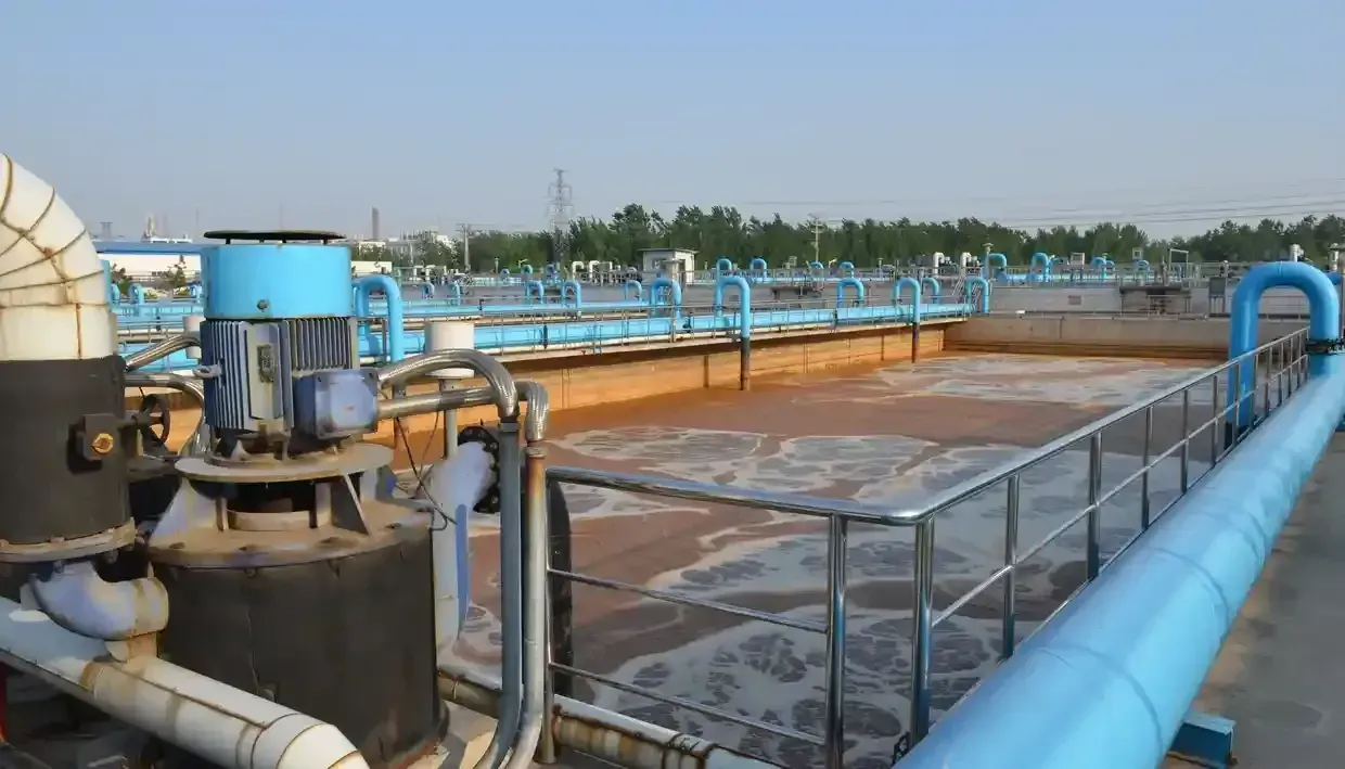 Industrial water treatment facility with blue pipes, pumps, and a large concrete tank under clear sky