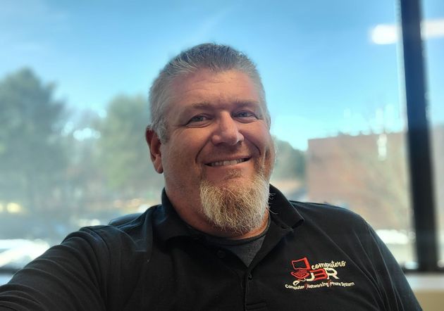 A smiling person with a goatee wearing a black polo shirt with a logo, photographed indoors with windows in the background.
