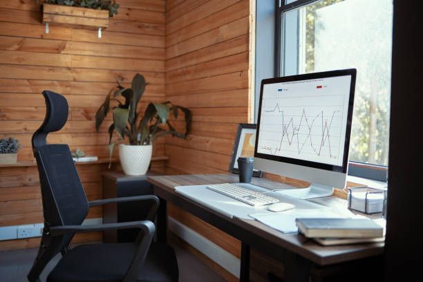 Home office desk with computer monitor, chair, and potted plants by a window and wooden wall.