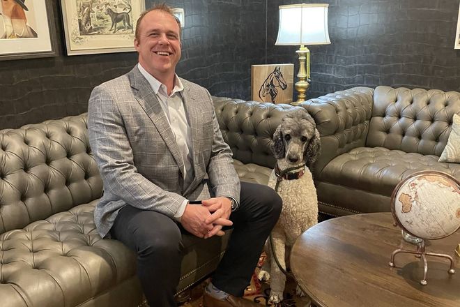 Man in blazer sits with a poodle on a tufted leather sofa in a room with decorative items.