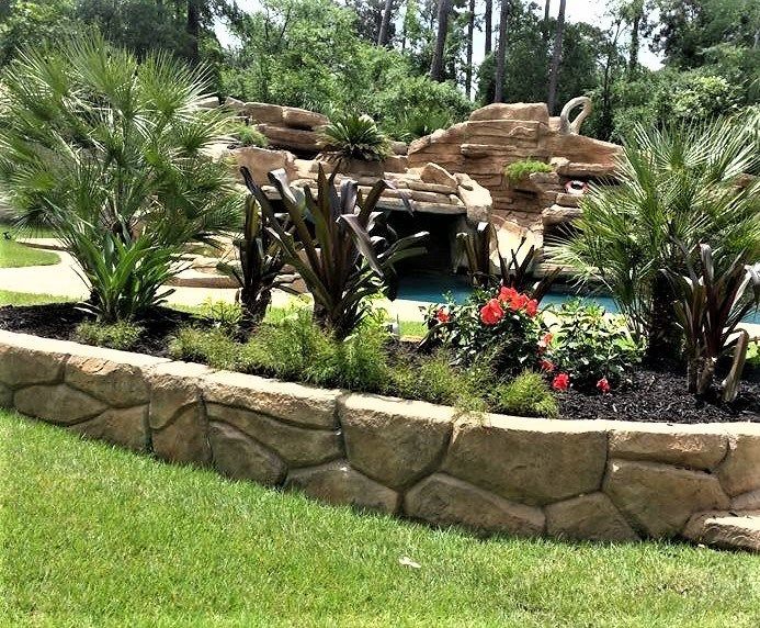 Stone-edged garden bed with palms and red flowers next to a lawn and rock waterfall.