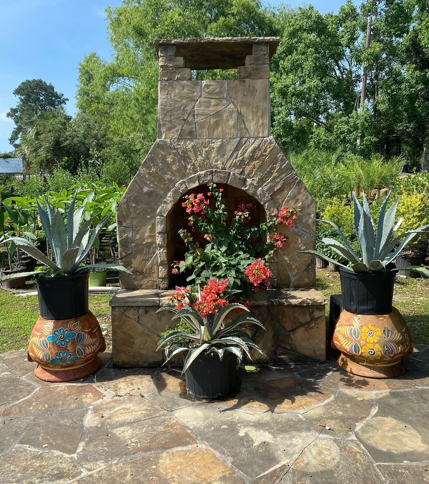 Stone fireplace with potted agave plants and colorful pots on a stone patio in a garden setting.