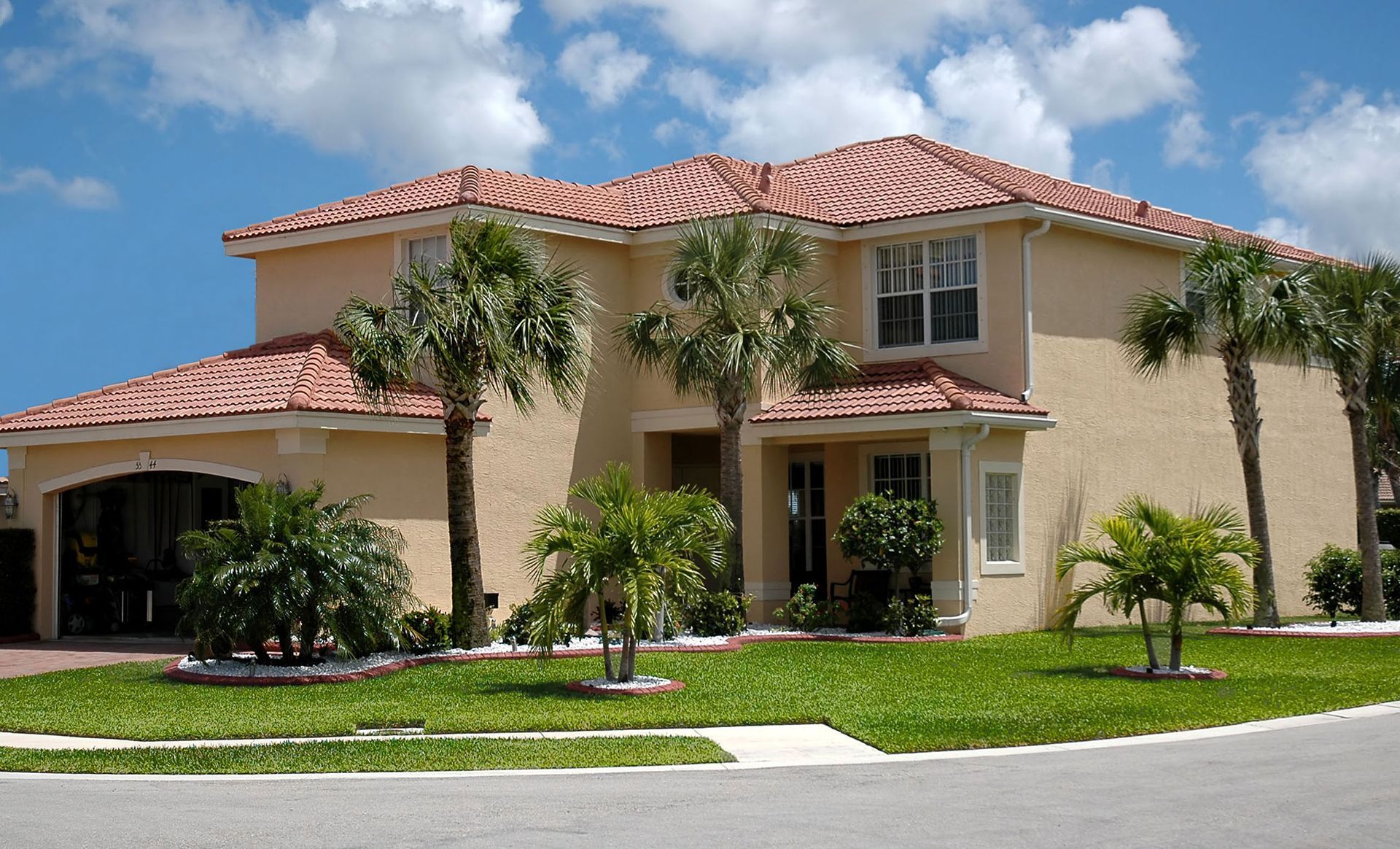 Two-story stucco house with red-tiled roof, palm trees, and green lawn under a blue sky.