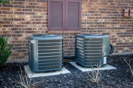 Two outdoor air conditioner units beside a brick house wall and window vents