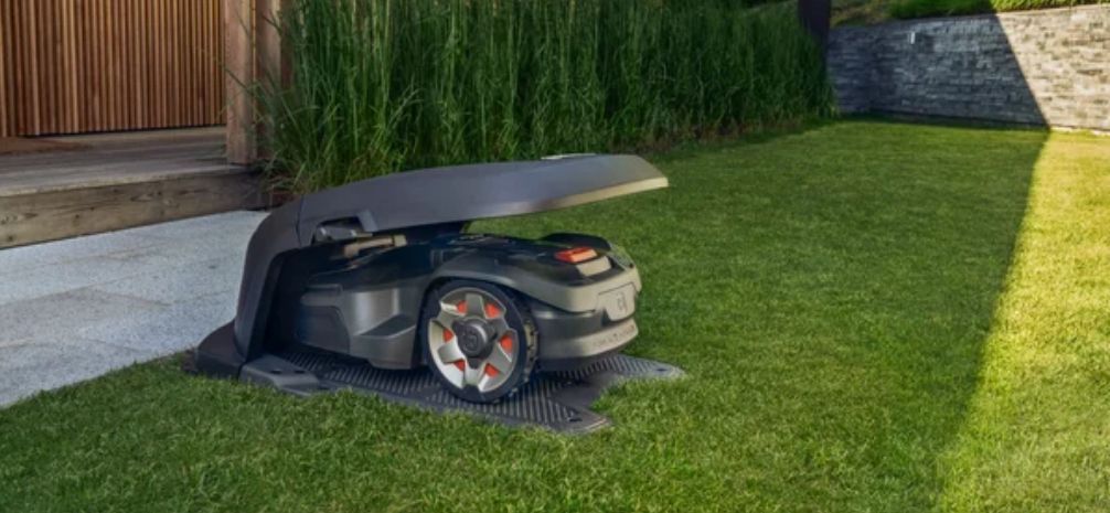 A robotic lawnmower sits in its charging station on a green lawn, near a wooden fence.