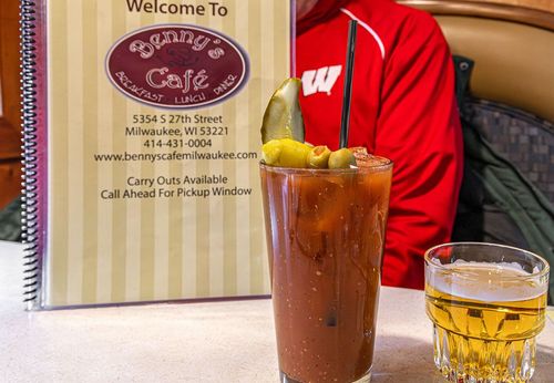 Cocktail and whiskey on a bar table beside a cafe menu, with a person in a red jacket behind them