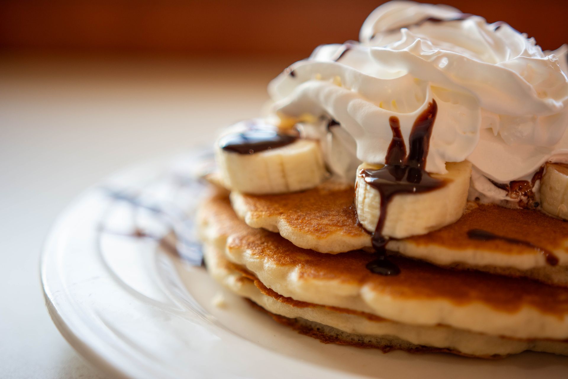 Stack of pancakes topped with banana slices, chocolate drizzle, and whipped cream on a white plate