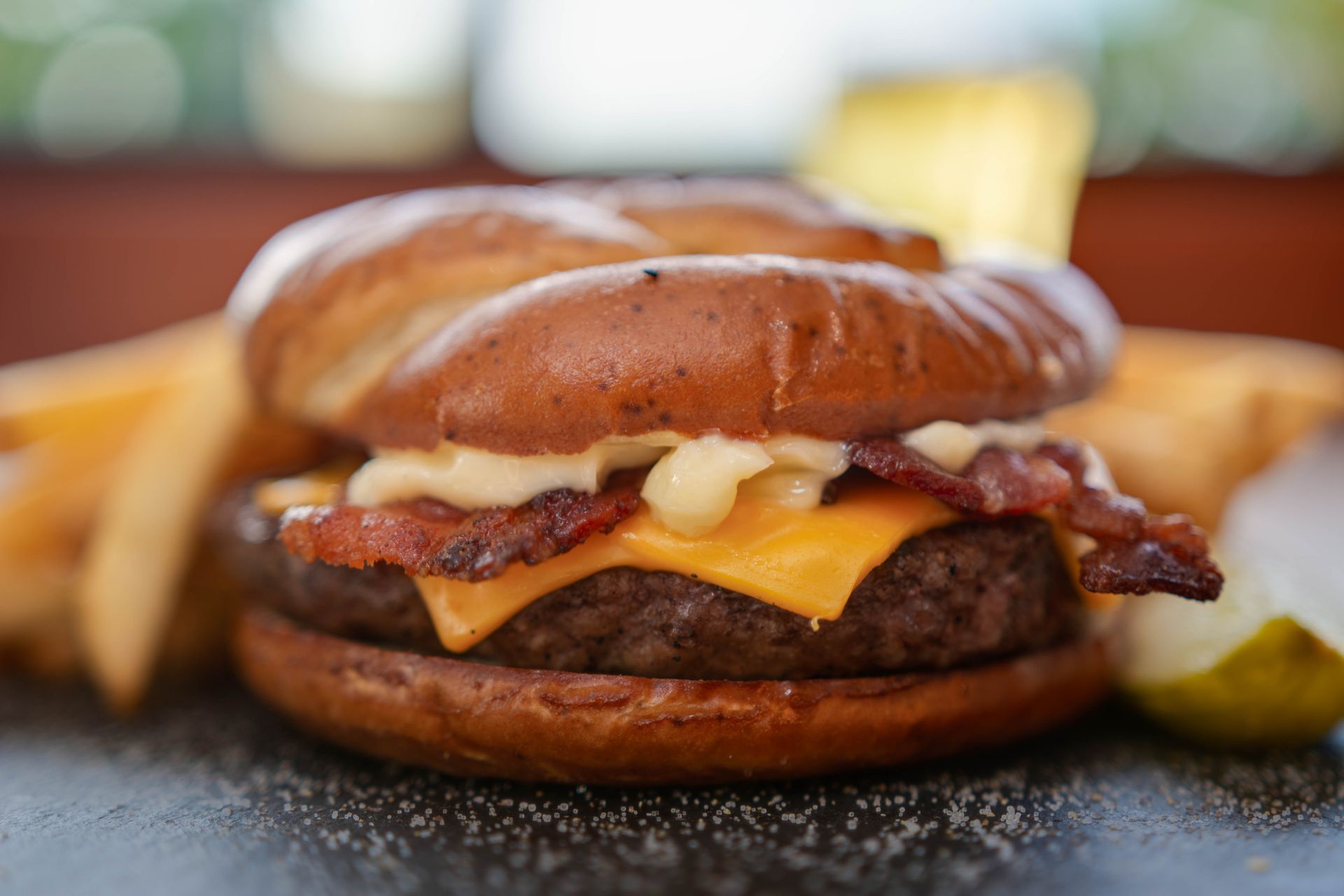 Close-up of a bacon cheeseburger with melted cheese on a bun, with fries and a pickle in the background