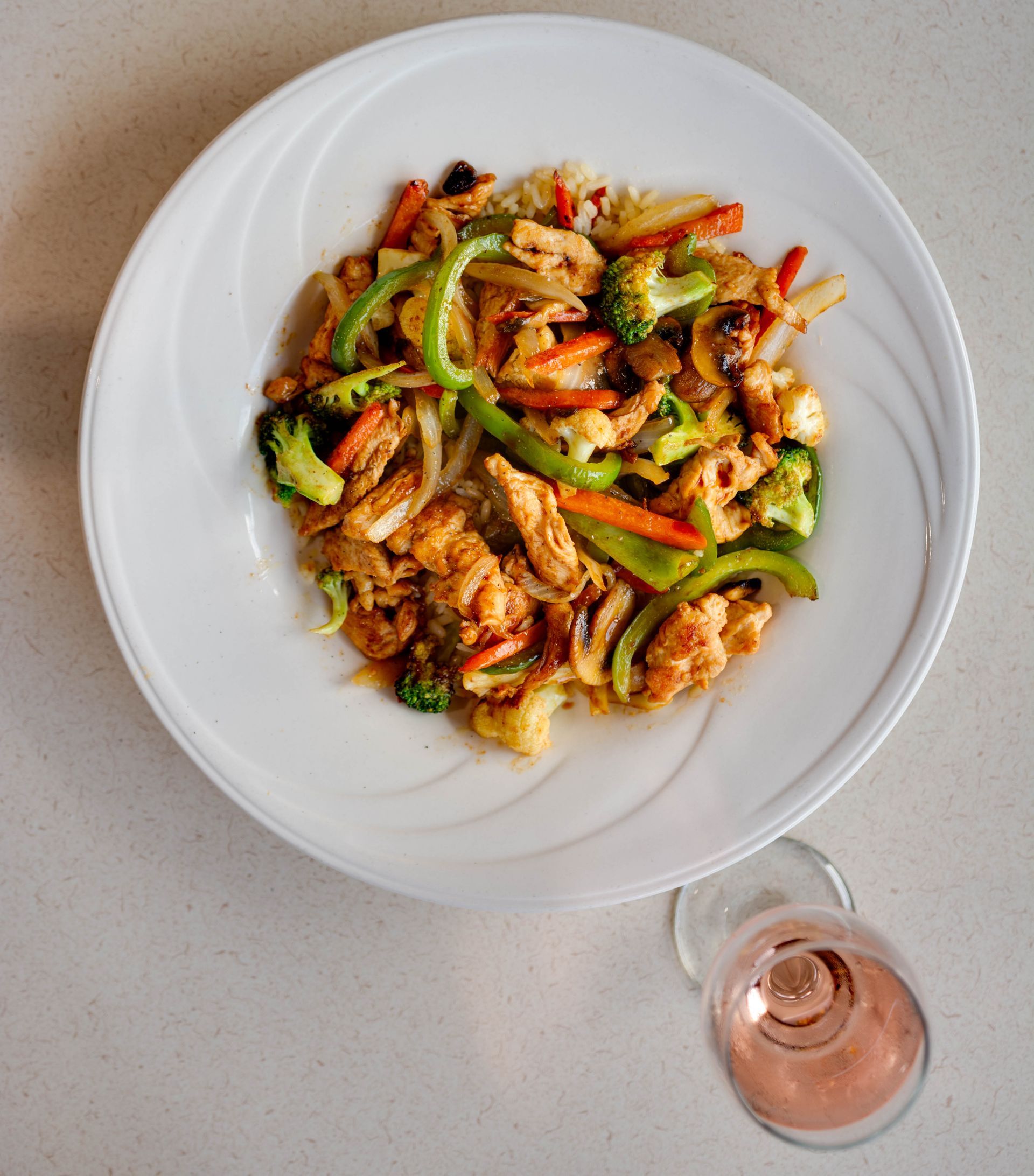 Plated stir-fry with vegetables and chicken on a white dish, beside a glass of rosé on a light table.