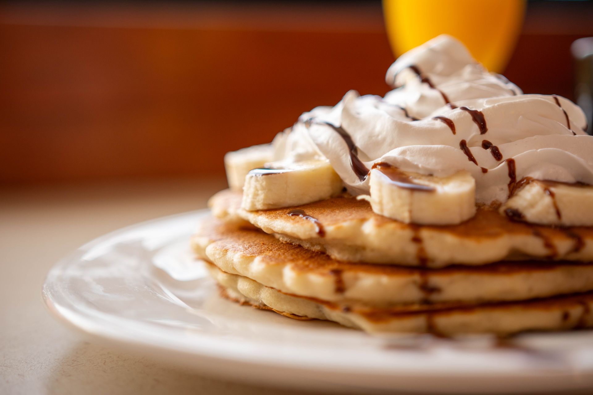 Stack of pancakes topped with banana slices, whipped cream, and chocolate drizzle on a white plate