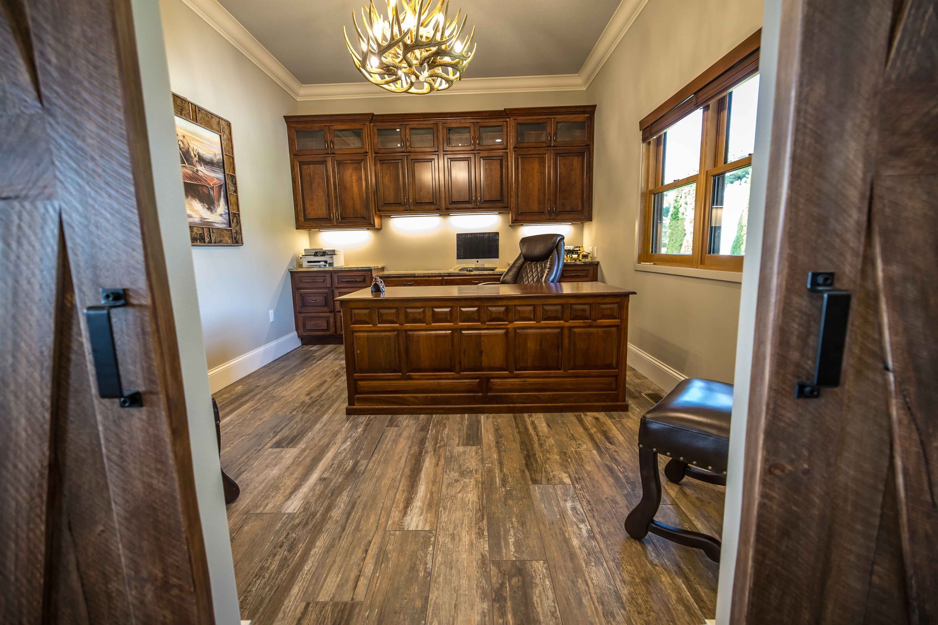A wood-paneled office with a large desk, cabinets, and a chandelier, seen through barn doors.