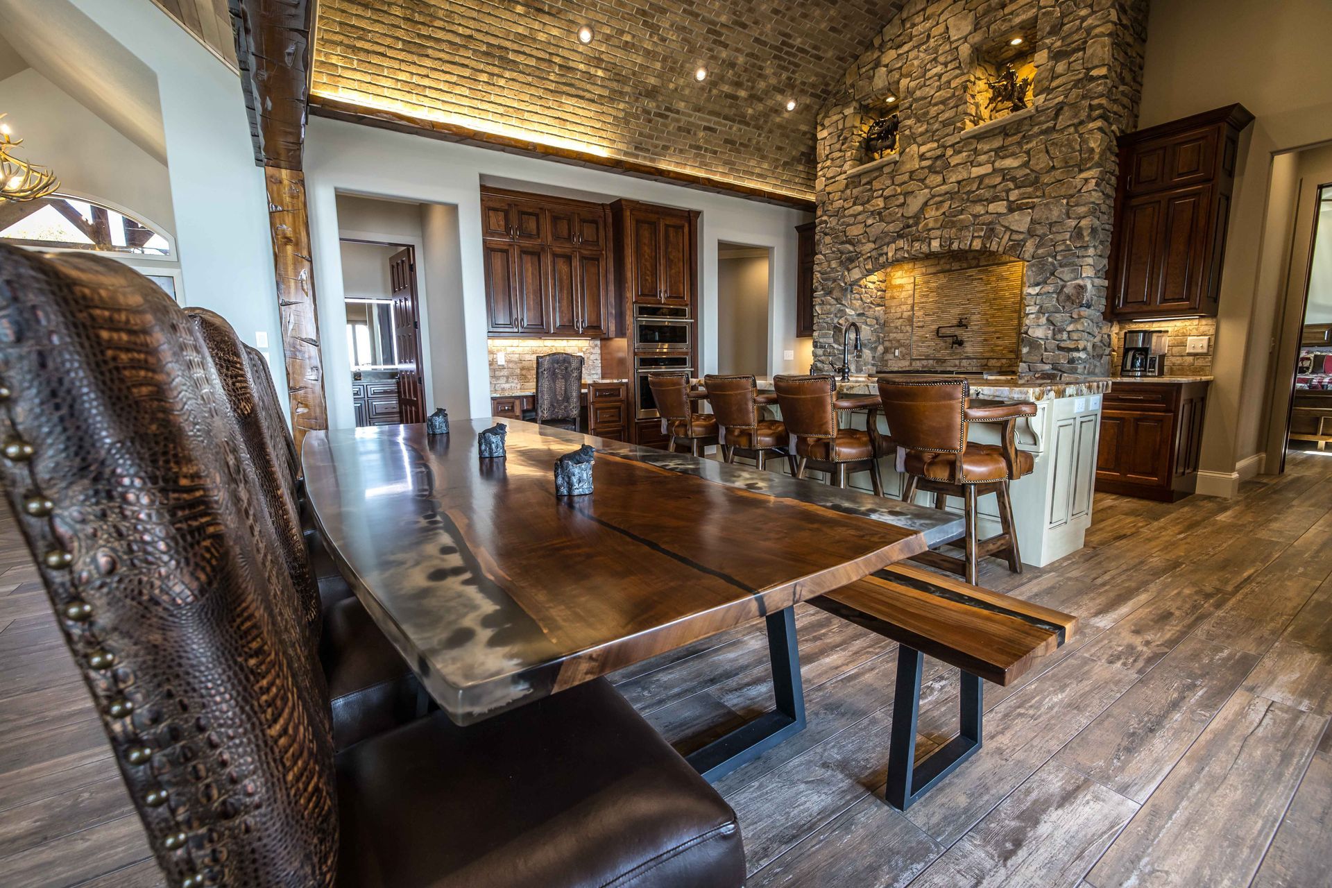 Dining room with a long wooden table and chairs, a stone wall, and a kitchen.