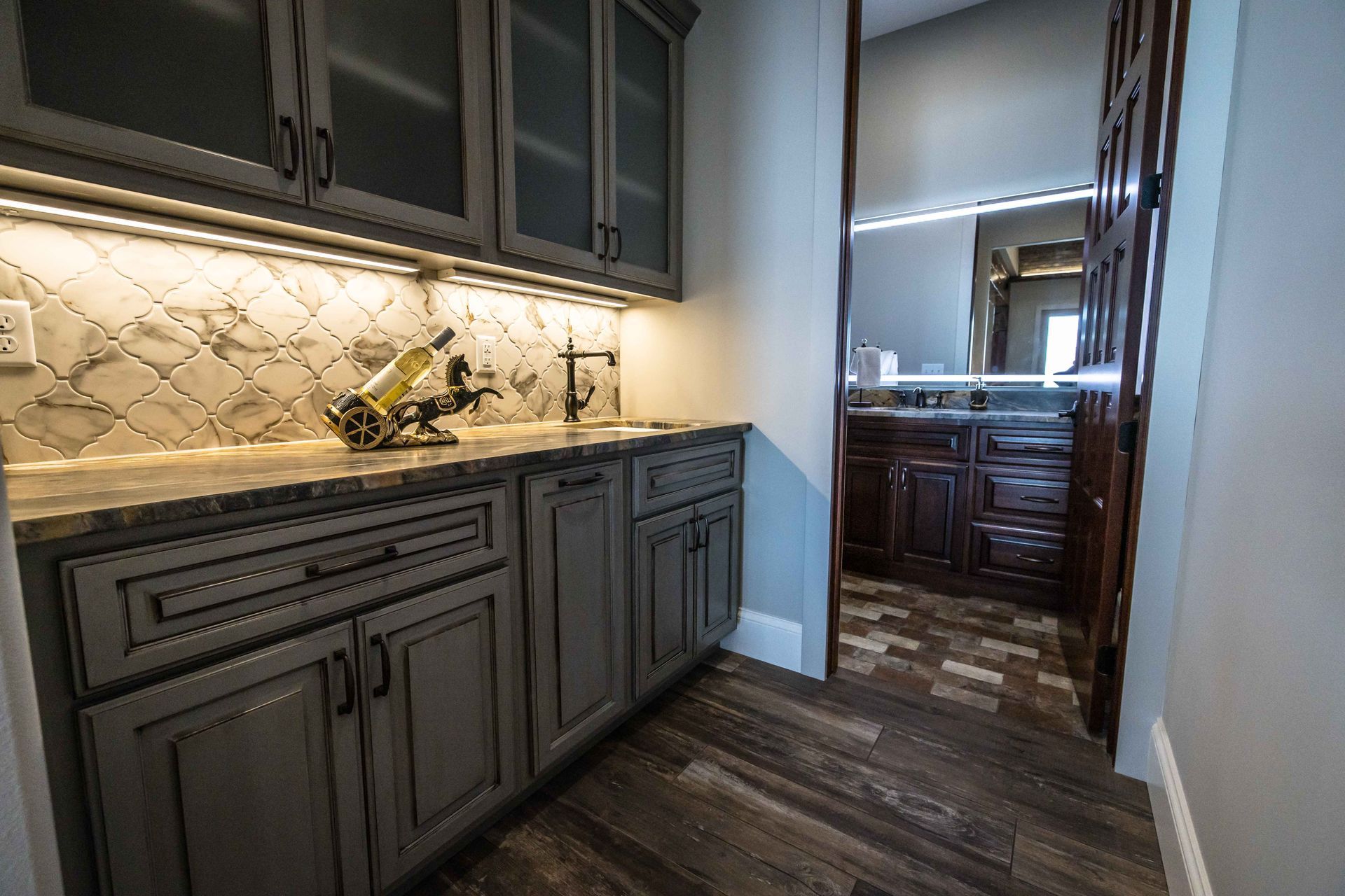 Gray cabinets with countertop, lighted backsplash, and an open doorway to a bathroom.