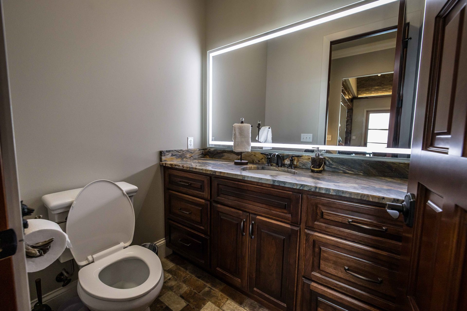 Bathroom with dark wooden vanity, granite countertop, lit mirror, and open toilet seat.