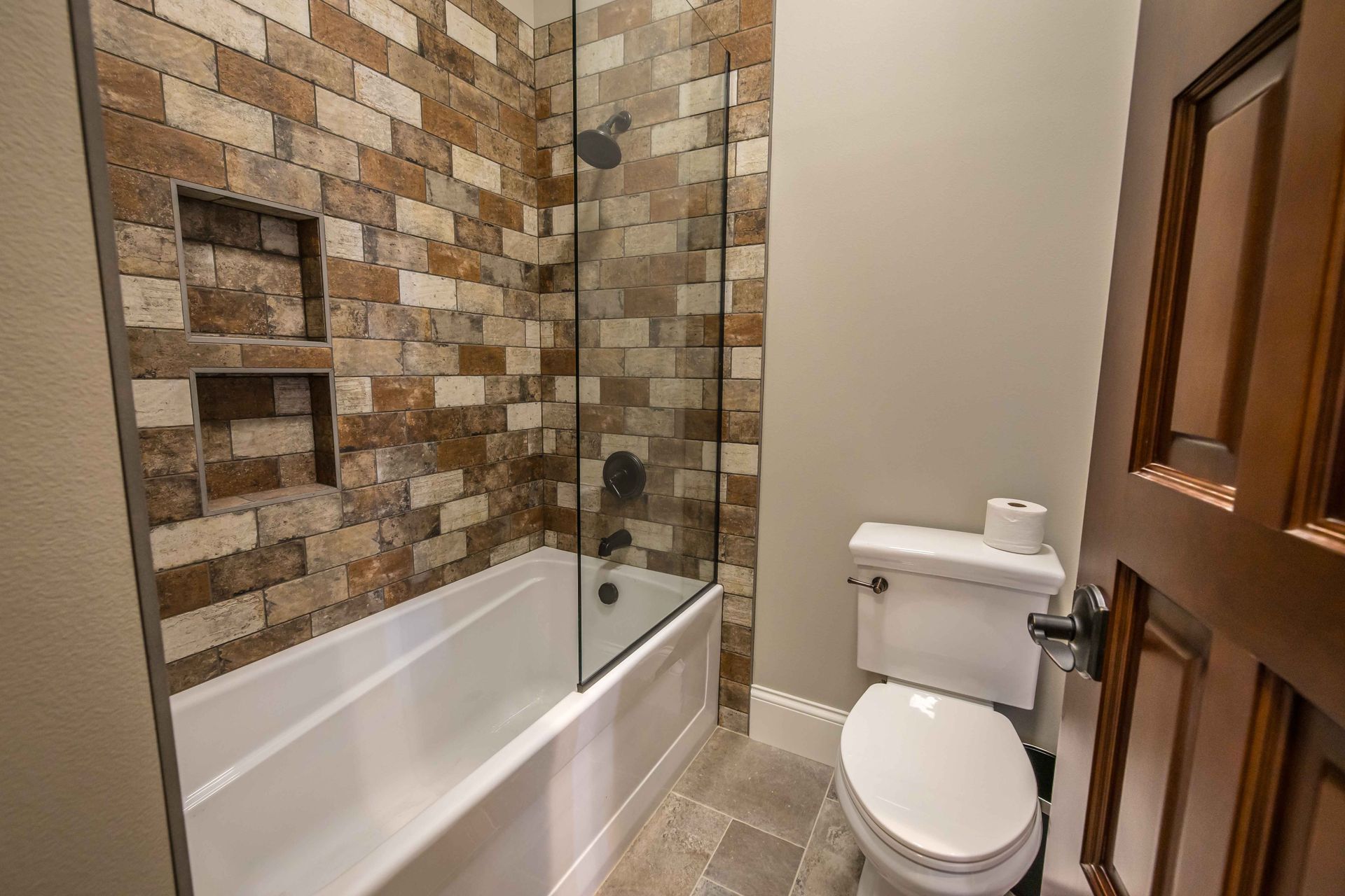 Bathroom with stone-tiled shower, white bathtub, toilet, and brown wooden door.