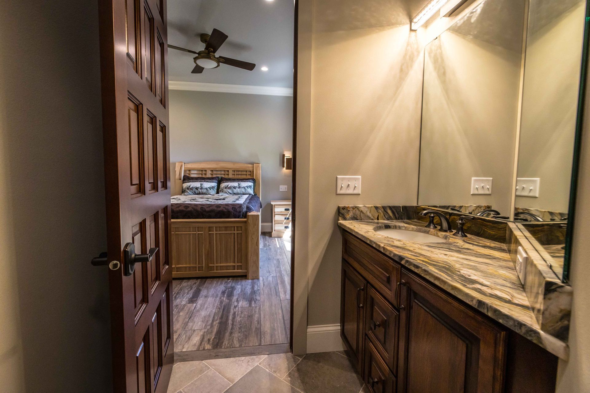 Bathroom doorway open to bedroom, vanity with mirror, wood door, and bed frame in view.