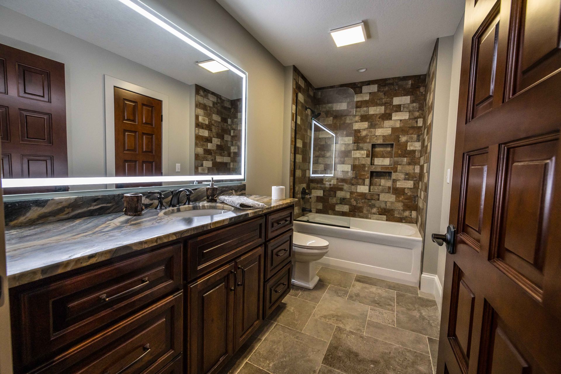 Bathroom with brown cabinetry, marble countertop, stone tile, and a bathtub.