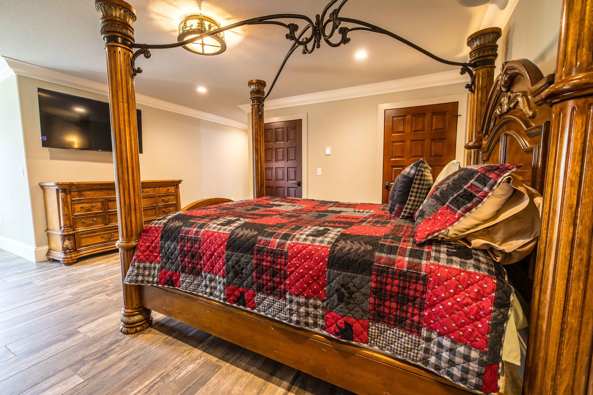 Bedroom with large wooden four-poster bed, plaid quilt, wooden dresser, TV on wall, and wooden doors.