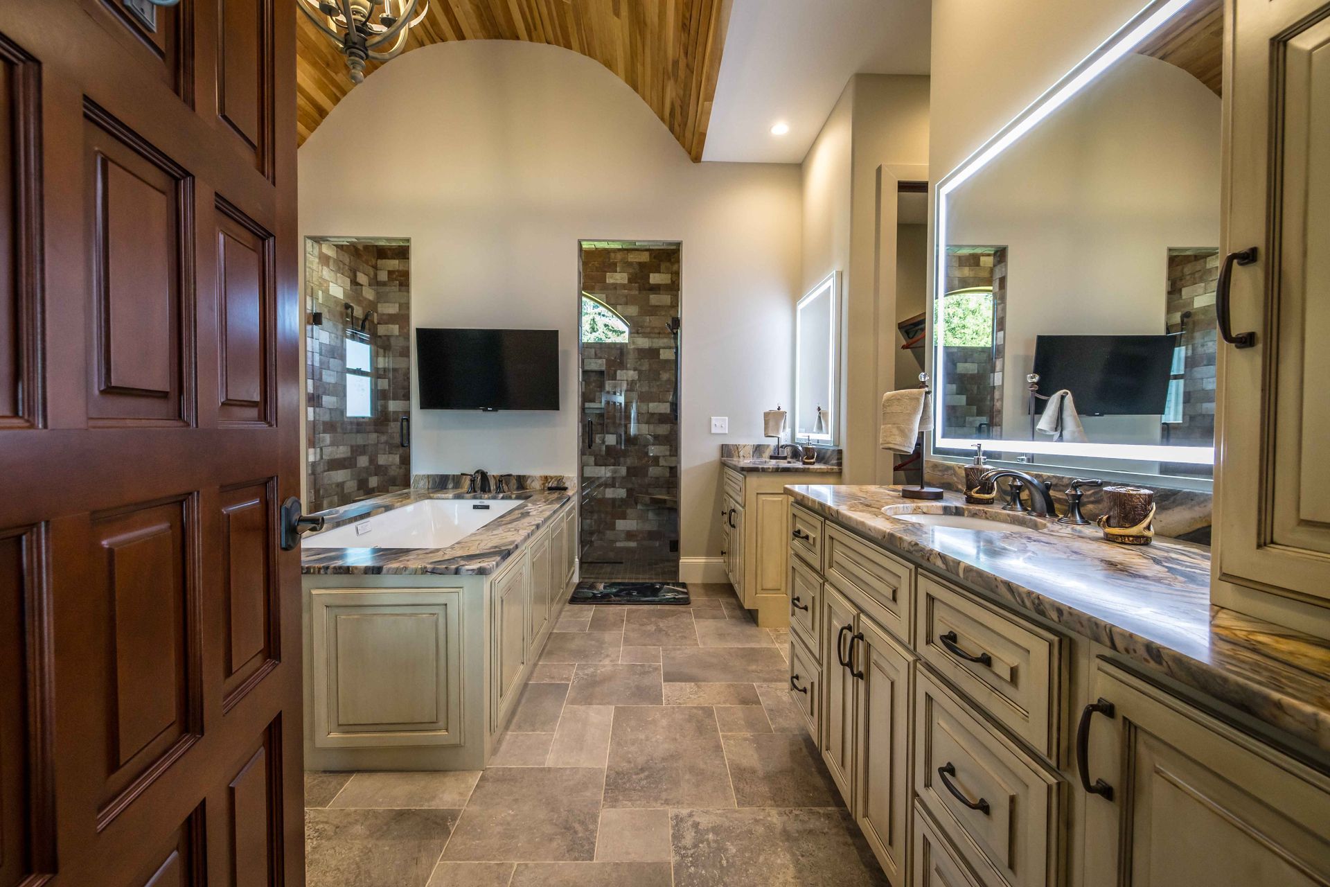 Bathroom with arched ceiling, stone tile, tan cabinetry, soaking tub, and television.