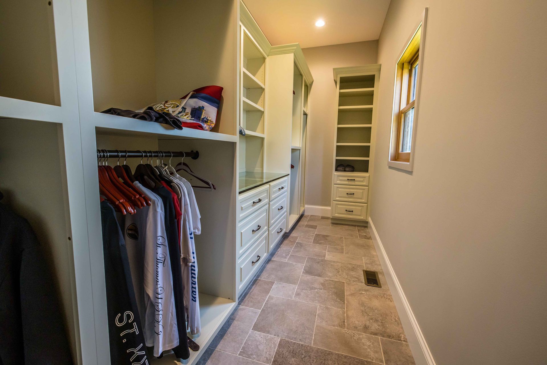 Walk-in closet with beige cabinetry, built-in shelves and drawers, clothing on hangers, and stone tile floor.
