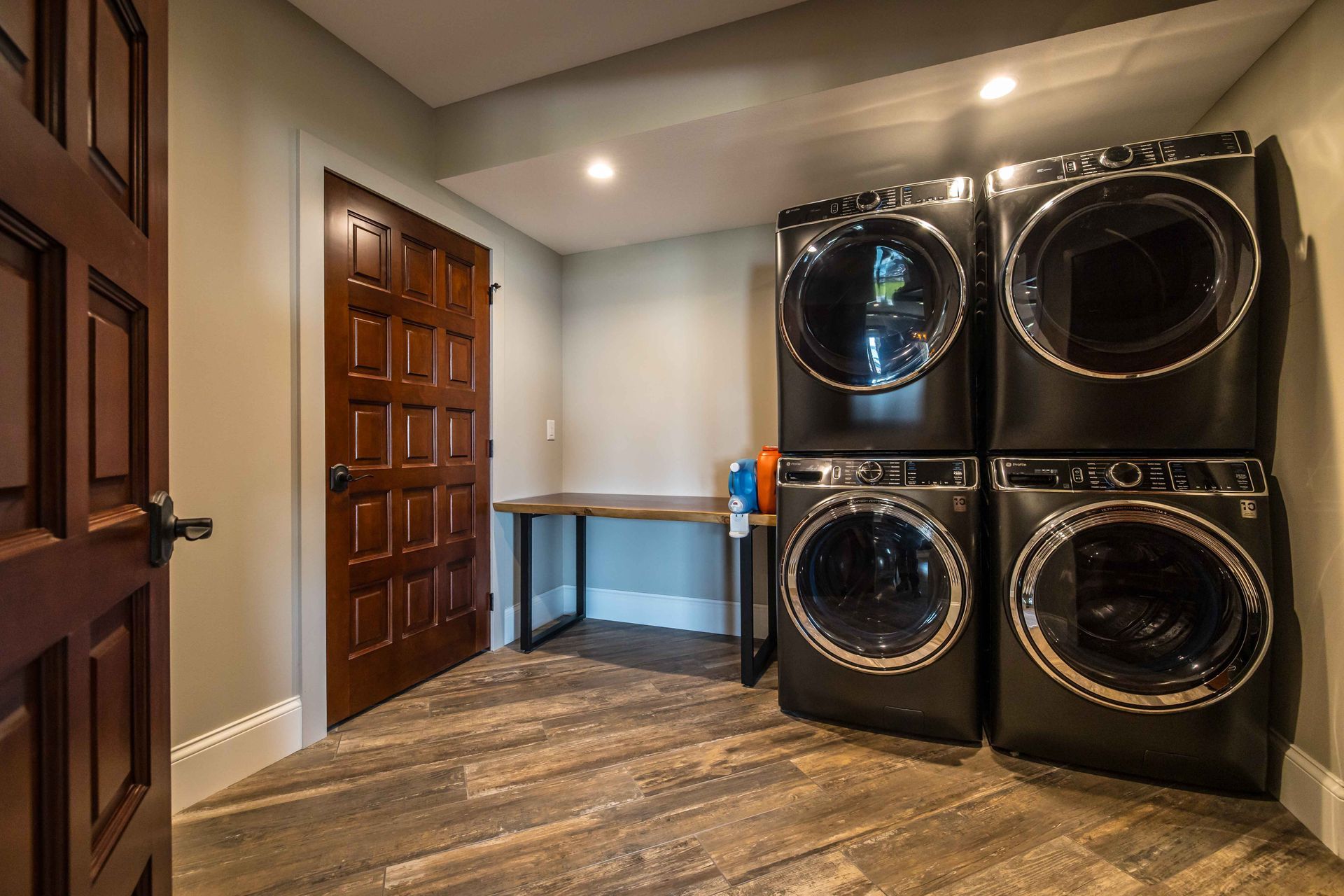 Laundry room with stacked black washer and dryer, wood door, and floor. Gray walls.