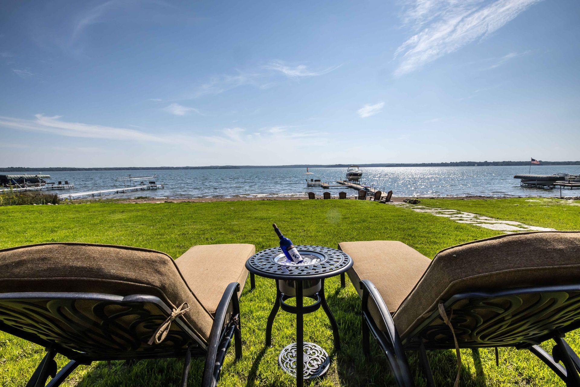 Two lounge chairs face water, with a small table holding a bottle in ice, on a grassy area under a blue sky.